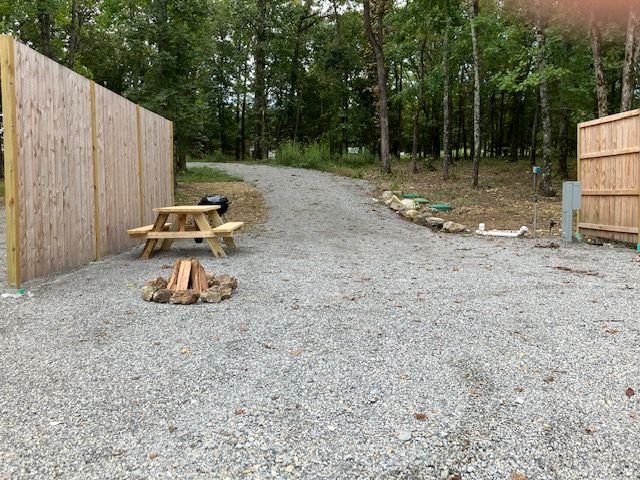 Gravel campsite with a wooden fence, picnic table, fire pit, and grill. Driveway leads into wooded area.
