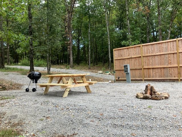 Camping site with picnic table, grill, and fire pit in gravel area, wooden fence and trees in background.