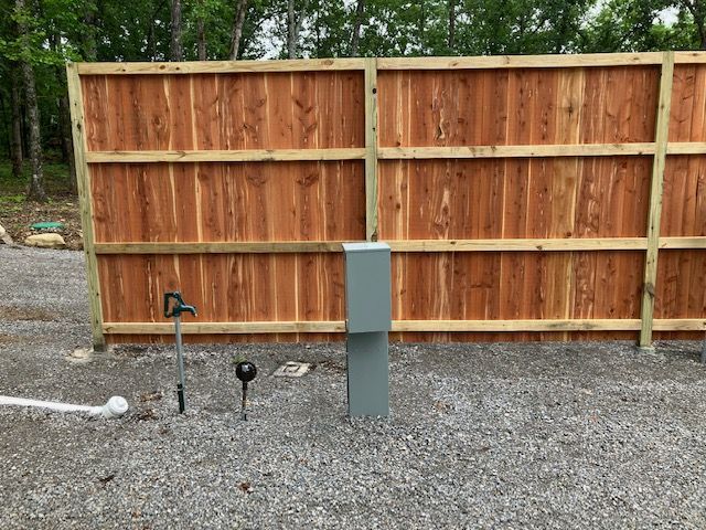 Wooden fence with a gray electrical box, water spigot, and gravel ground.