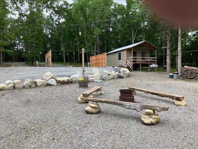Gravel campsite with a small cabin, fire pit, log benches, and surrounding trees.
