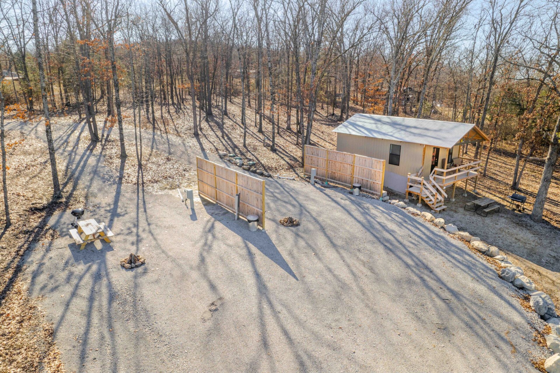Gravel driveway leading to a small cabin in a wooded area. Fencing on both sides of driveway. Bright sunny day.