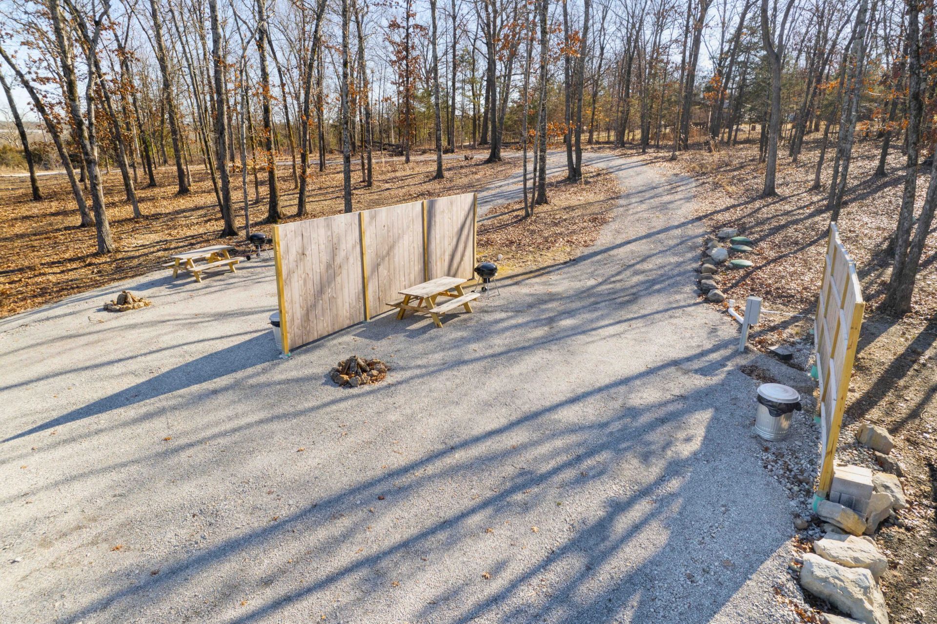Gravel driveway leading into a wooded area, bordered by rocks and wooden fences, with trees in the background.