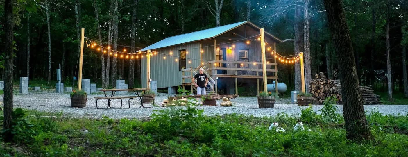 A cabin in the woods decorated with string lights, a person on the porch, and a fire pit.