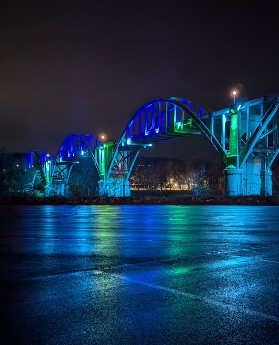 Blue and green illuminated bridge reflecting on water at night.