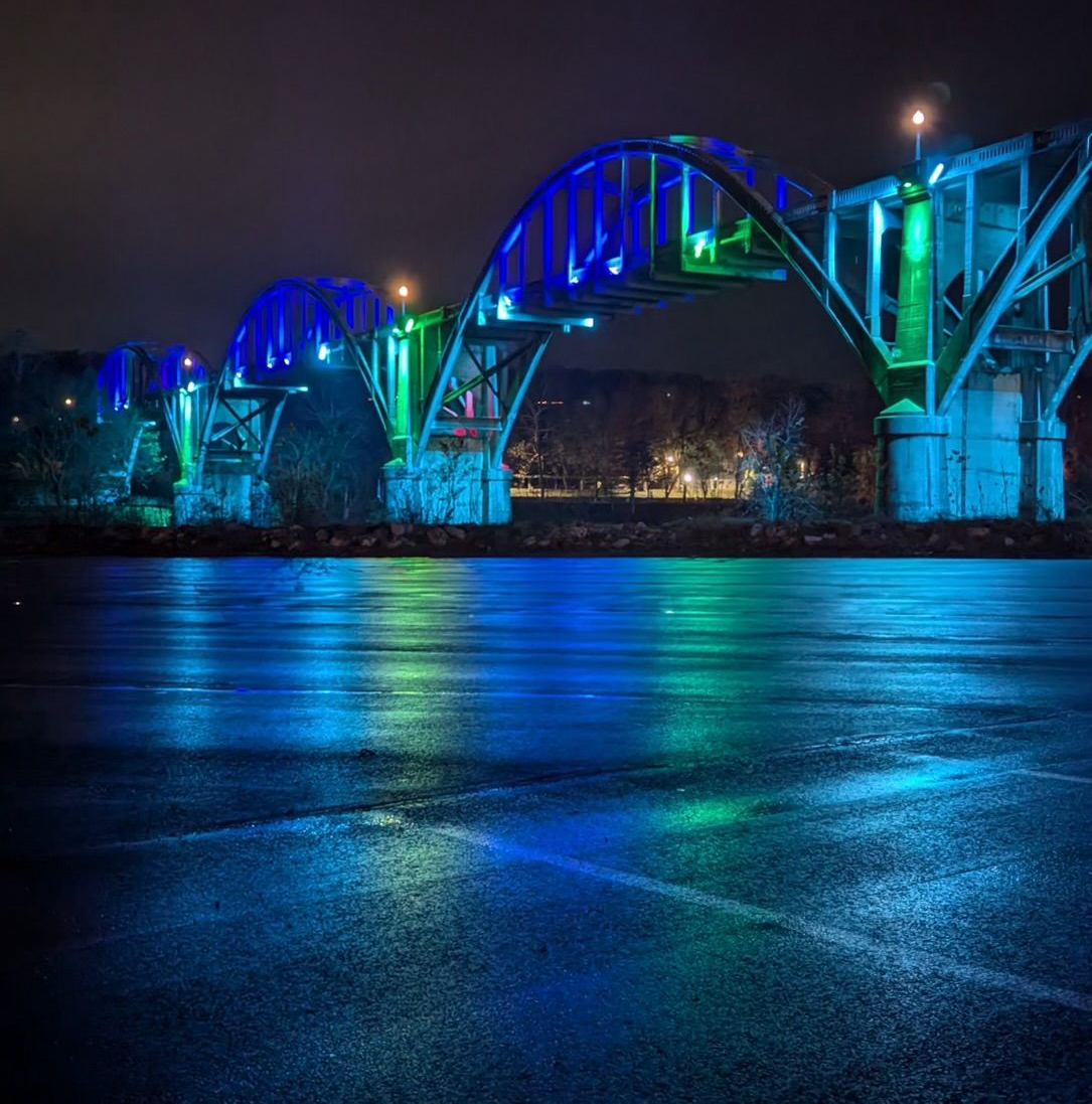 Nighttime view of a bridge lit with blue and green lights, reflected in the water below.
