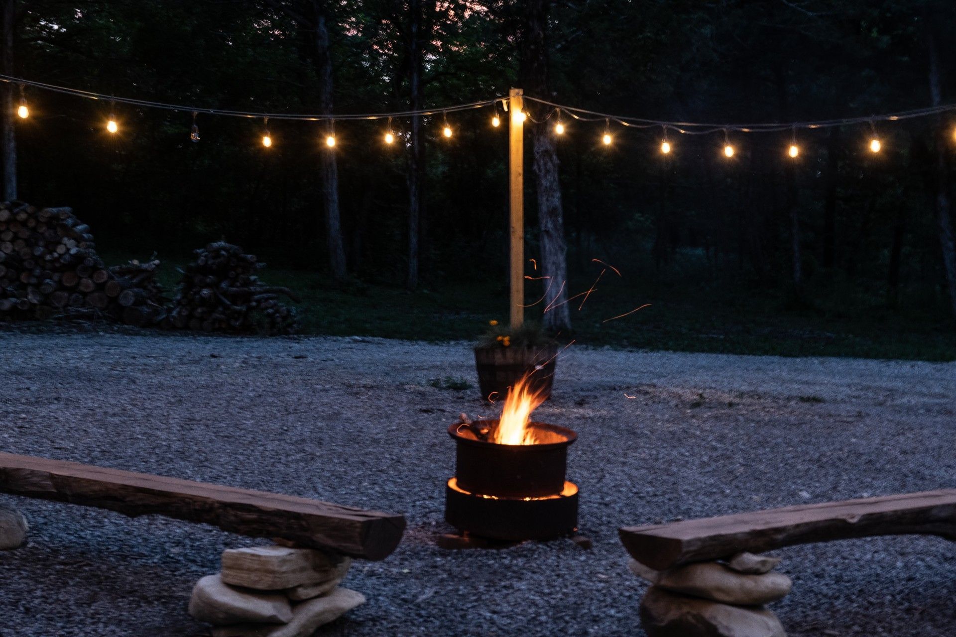 Campfire at dusk with string lights and rustic benches.