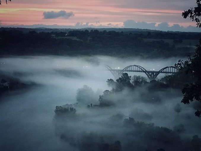 Foggy landscape with a bridge spanning over a river at dusk. Soft pink and blue hues fill the sky.