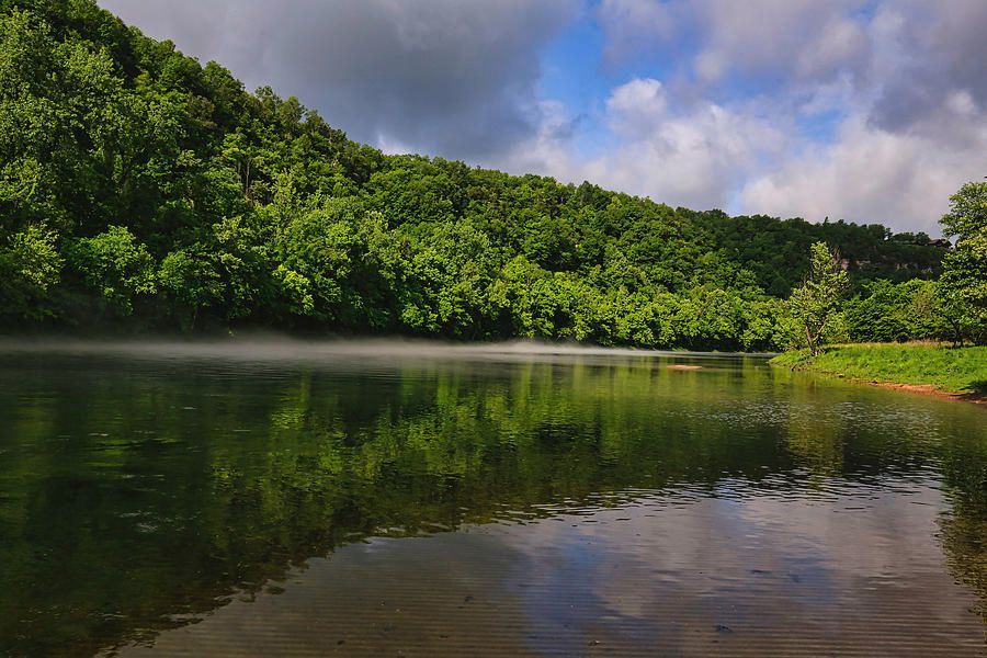 A river flows in front of a tree-covered hillside under a partly cloudy sky.