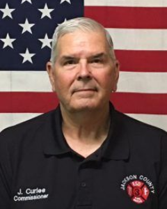 A man wearing a Jackson County Fire District 3 Commissioner polo shirt, standing in front of an American flag.