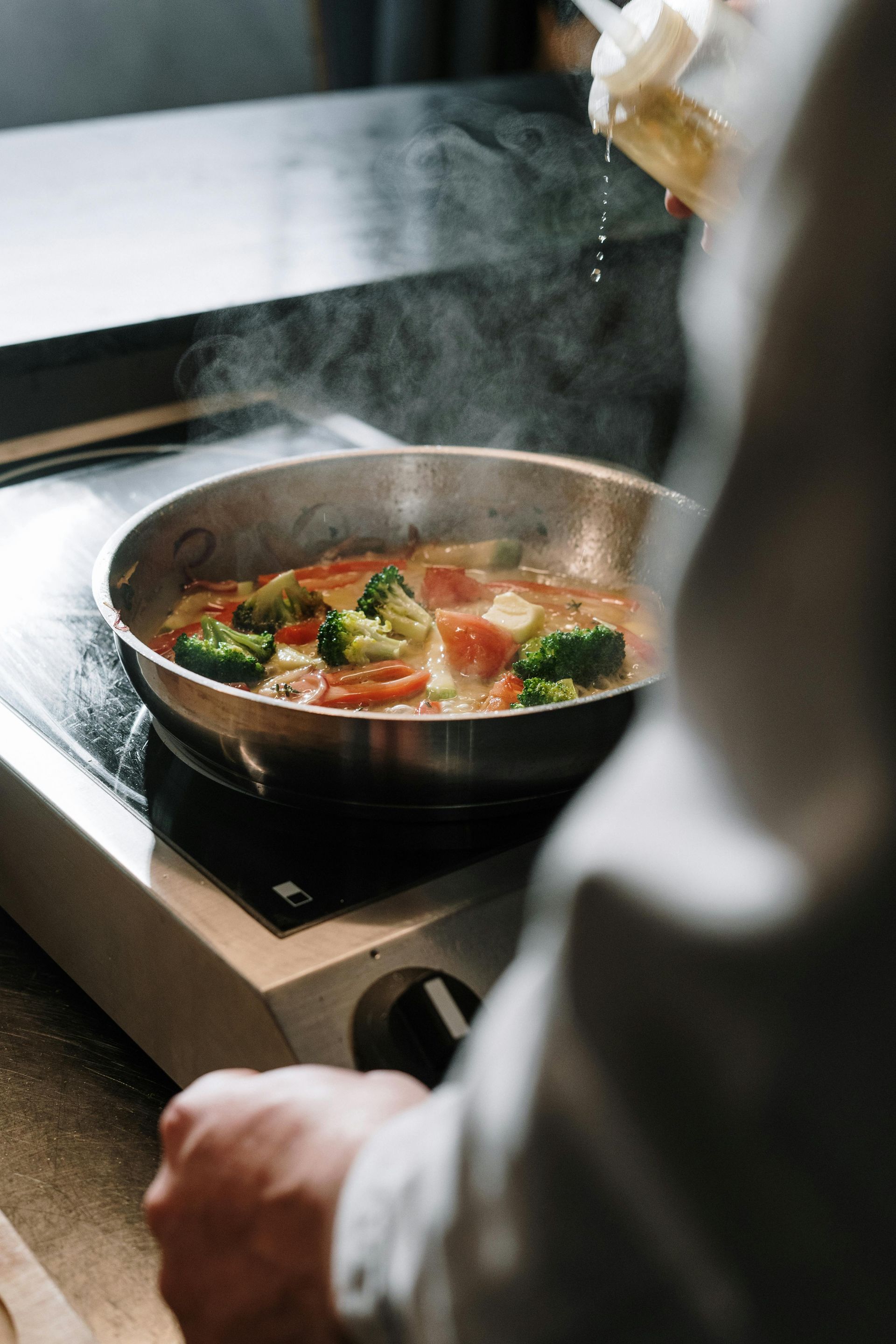 Chef seasoning vegetables in a pan on a stovetop, steam rising.