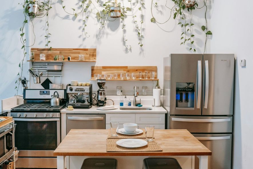 A small, modern kitchen with wooden accents and live plants. Features a stove, fridge, sink, and dining table.