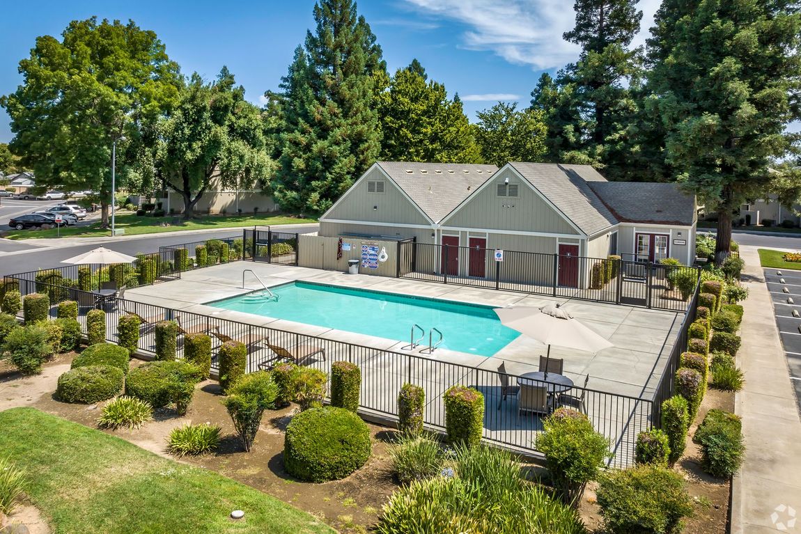 Pool with blue water surrounded by concrete, greenery, and residential buildings on a sunny day.