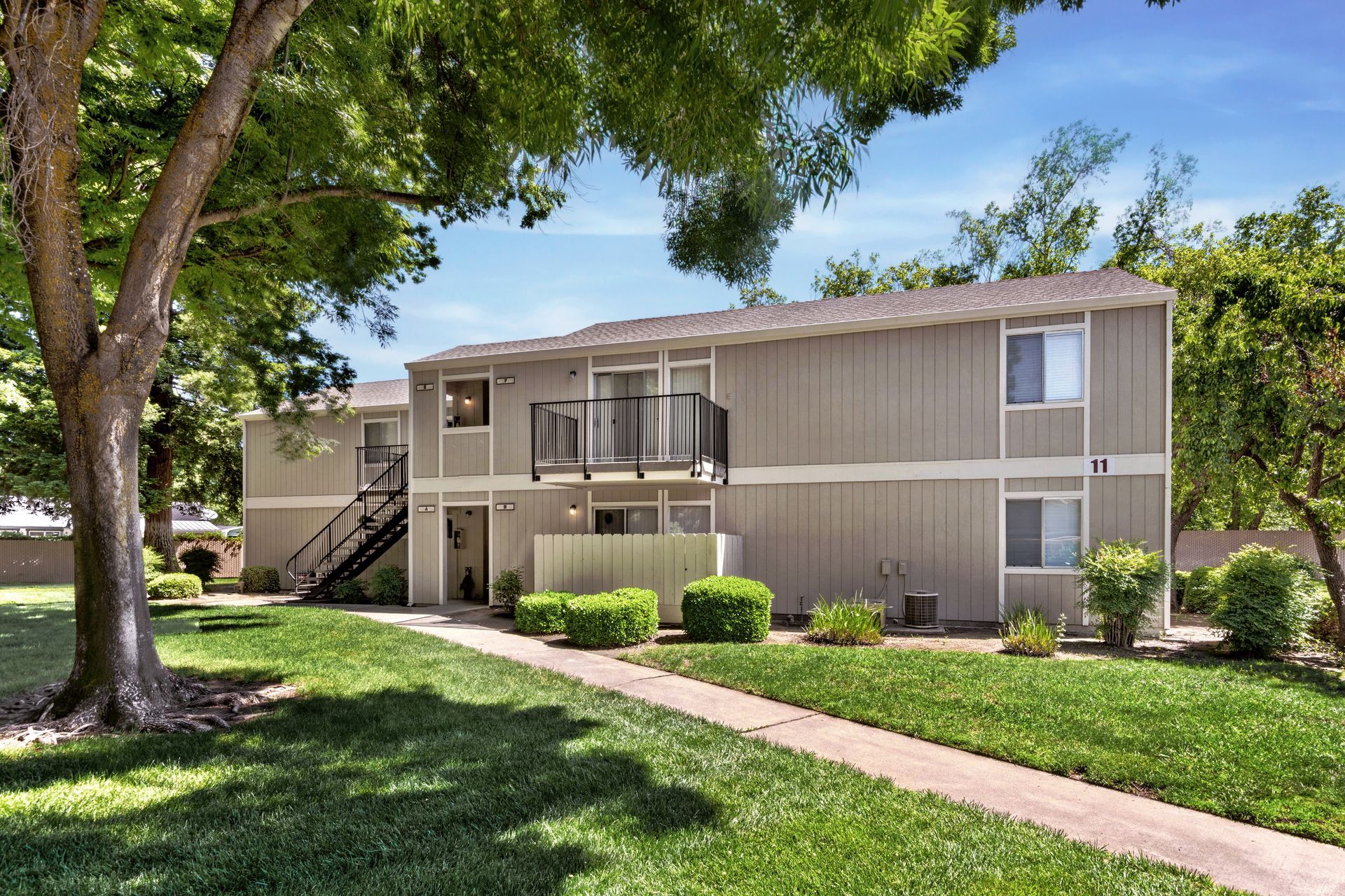 Two-story beige apartment building with a balcony, green grass, and a walkway under a blue sky.