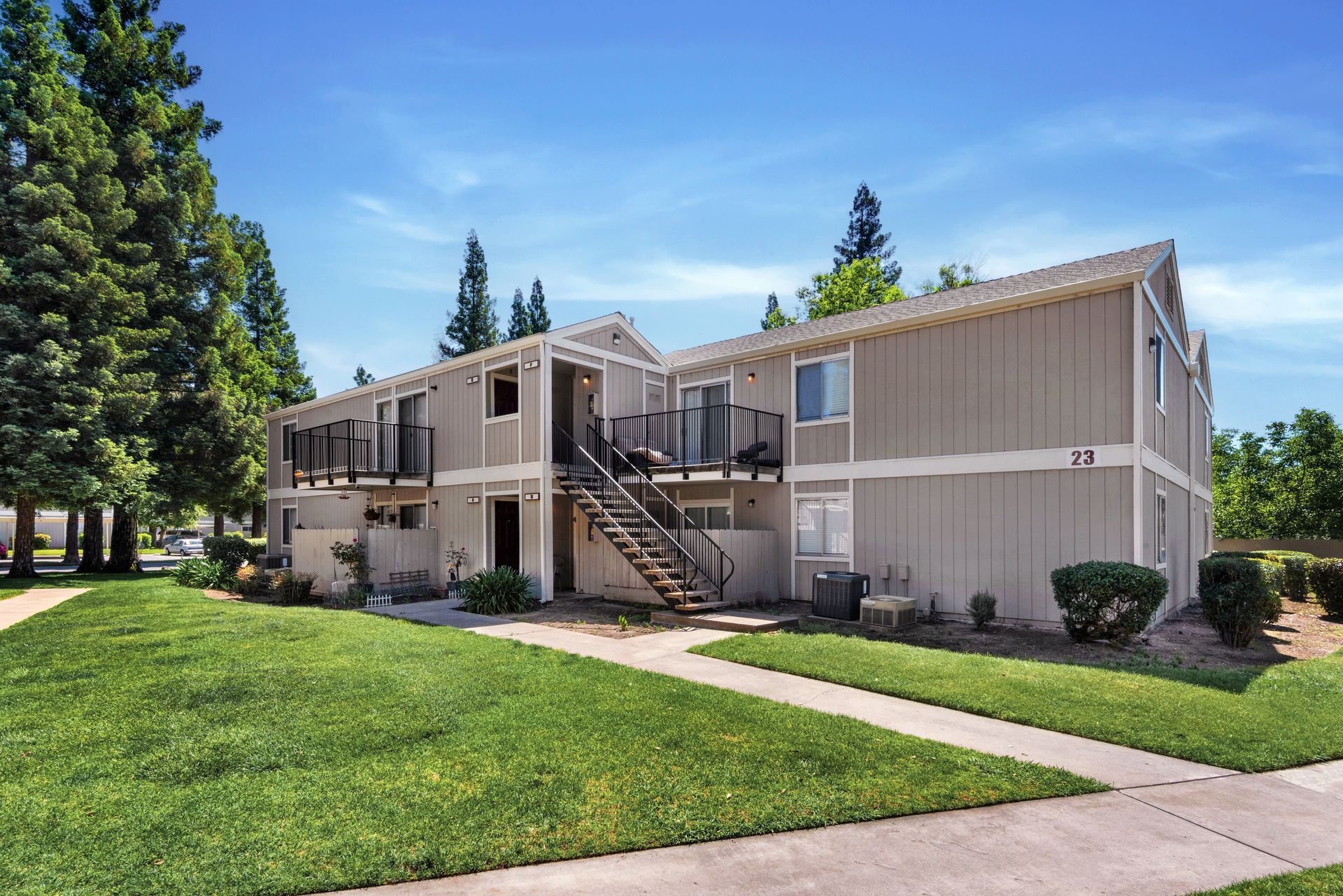Two-story apartment building with light-colored siding, stairs, and balconies on a green lawn under a blue sky.