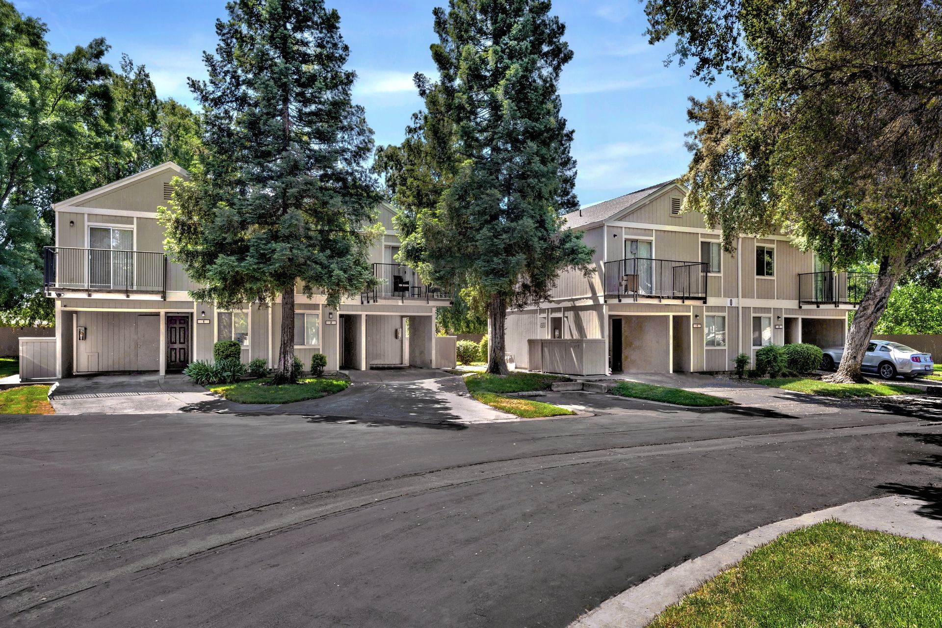 Two-story townhomes, light gray with balconies, on a paved road. Trees and greenery surround.