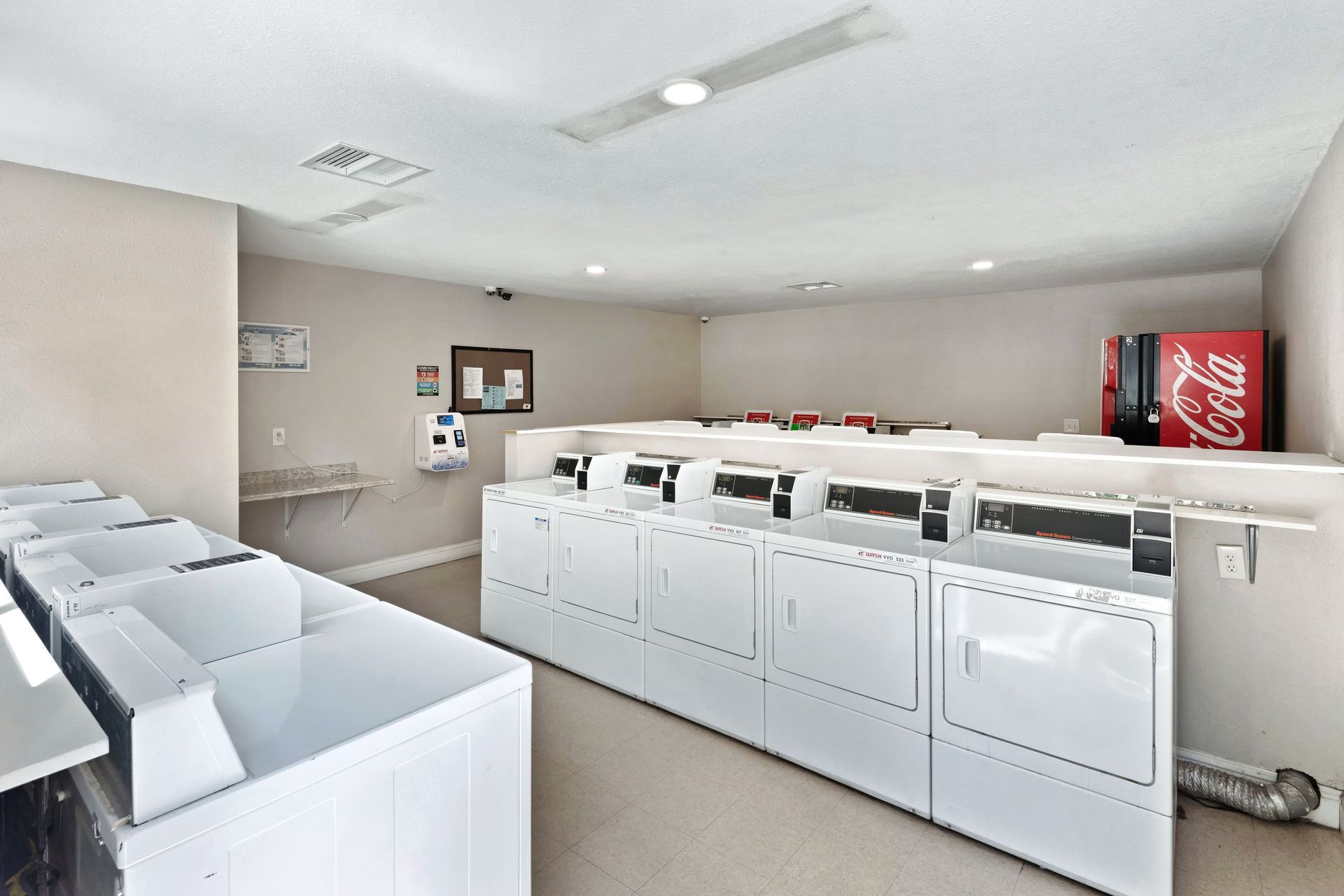 A well-lit laundromat with several white washing machines and dryers. A Coca-Cola vending machine is visible.