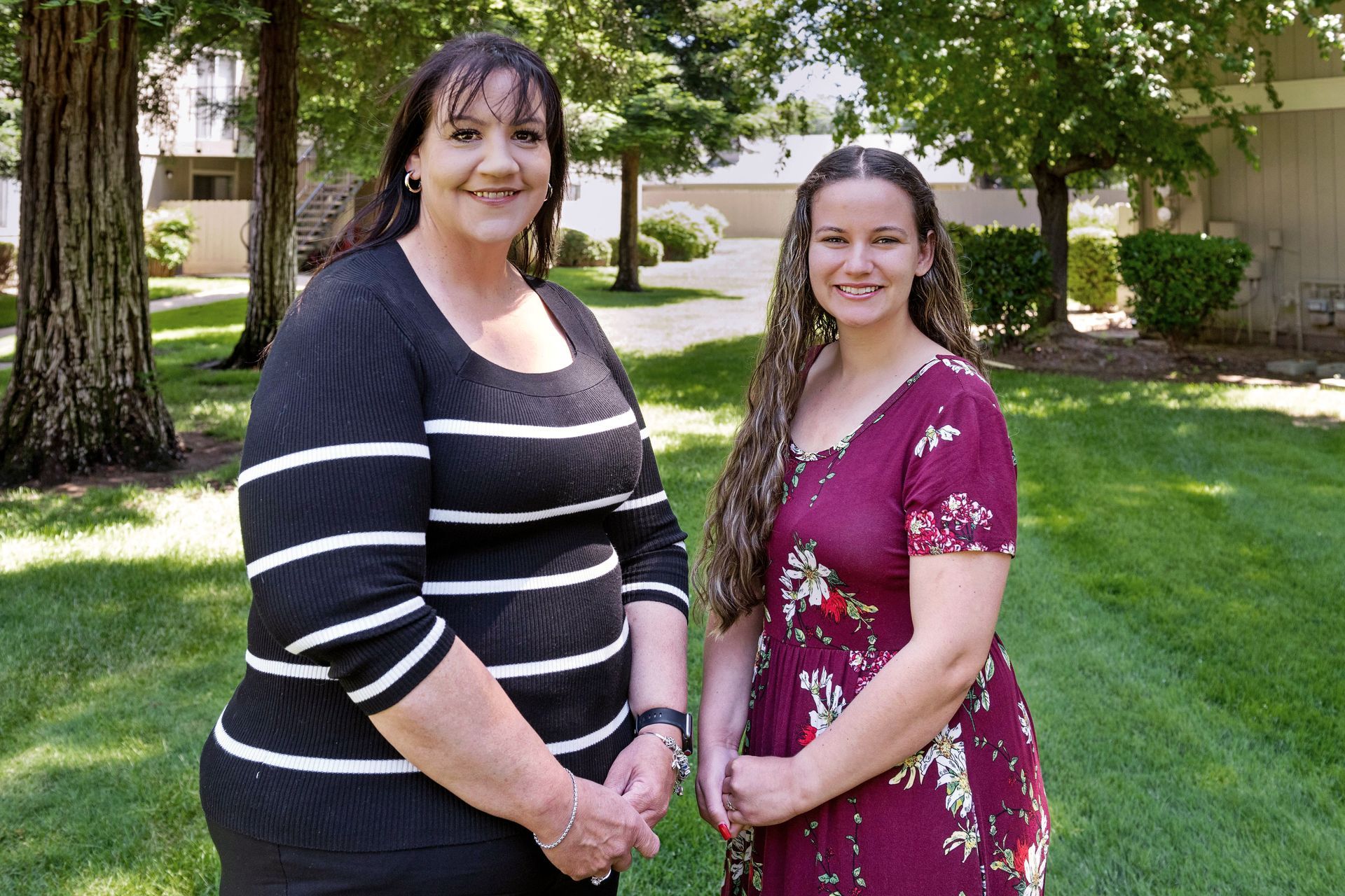 Two women smiling, standing on grass in front of a building and trees. One wears a striped shirt, the other a floral dress.