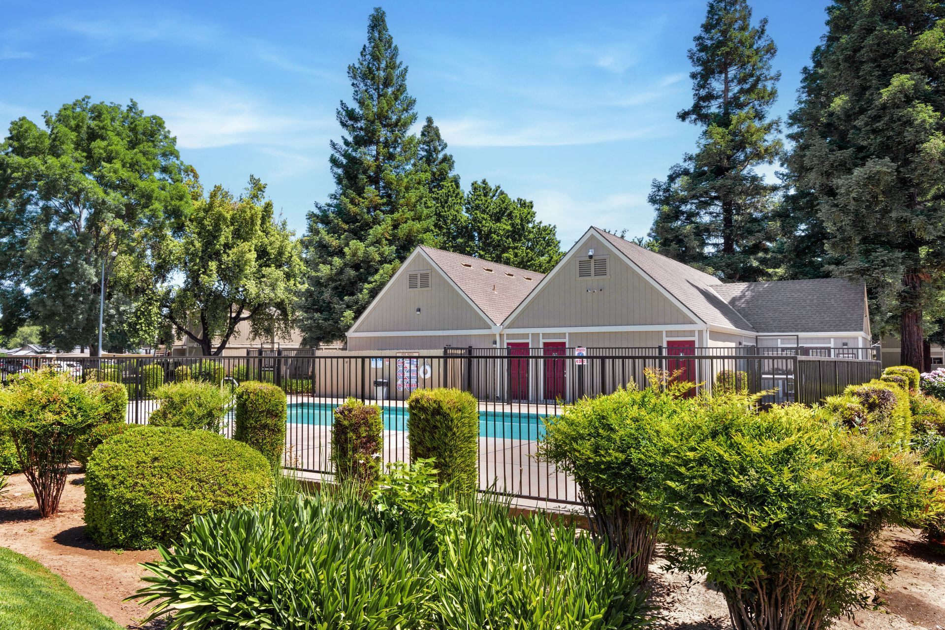Pool area with beige building, black fence, green bushes, and blue sky.