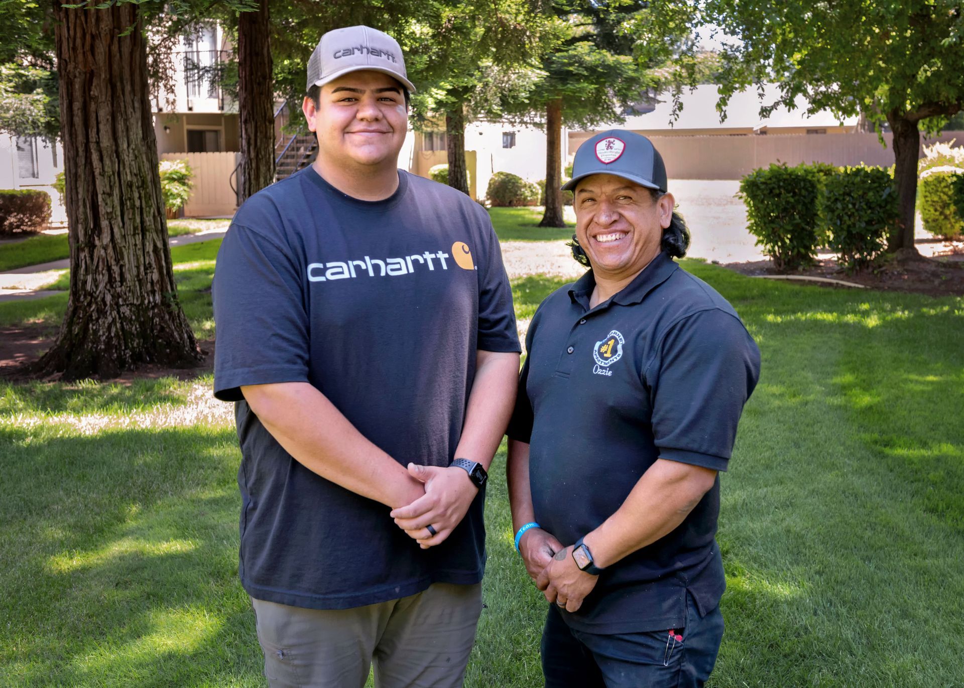 Two men standing in a park, one wearing a Carhartt shirt, the other a polo shirt, smiling.
