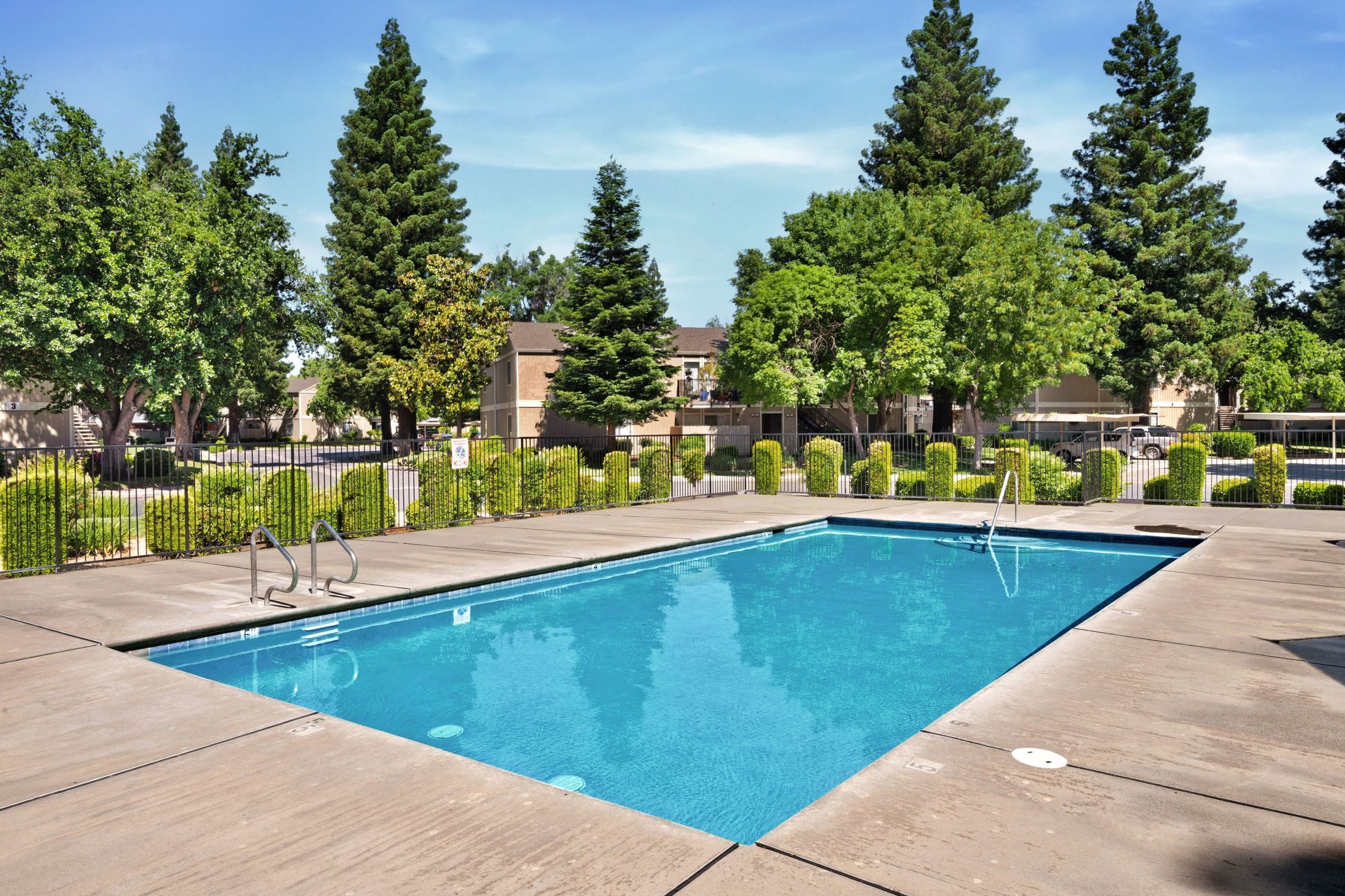 Swimming pool surrounded by a concrete deck, lounge chairs, and apartment buildings under a blue sky.