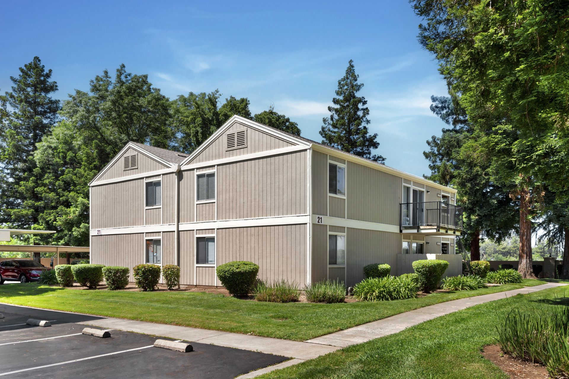 Two-story tan apartment building with green lawn, trees, and clear blue sky.