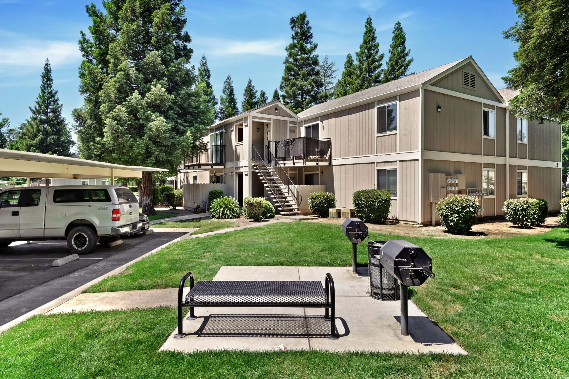 Apartment complex with two-story buildings, grass, grills, and a bench. A white truck is parked nearby.