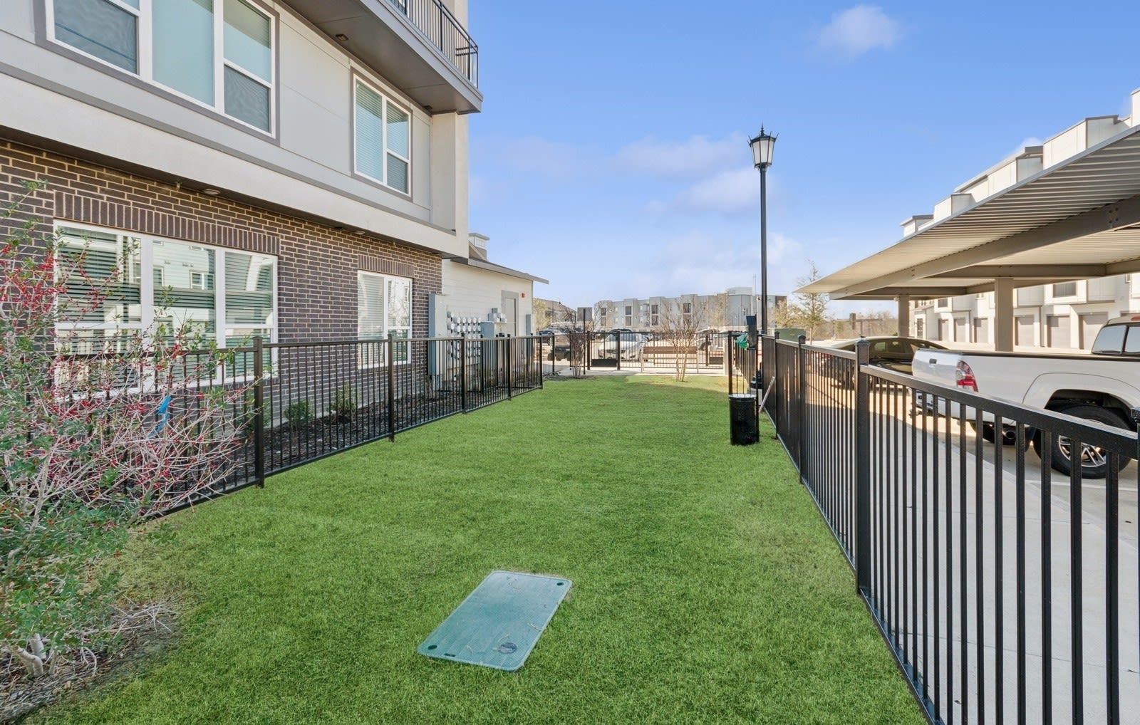Grassy communal courtyard with a black metal fence, adjacent apartment building, and a streetlamp.