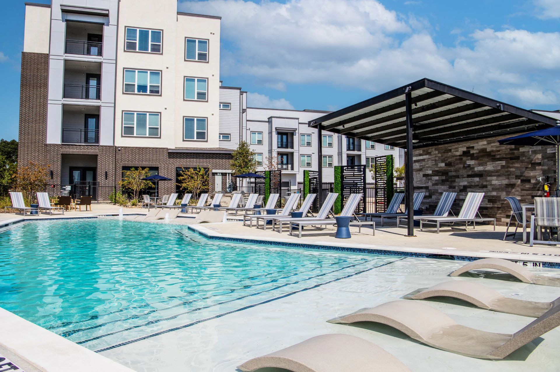 Swimming pool with lounge chairs, next to an apartment building under a blue sky.