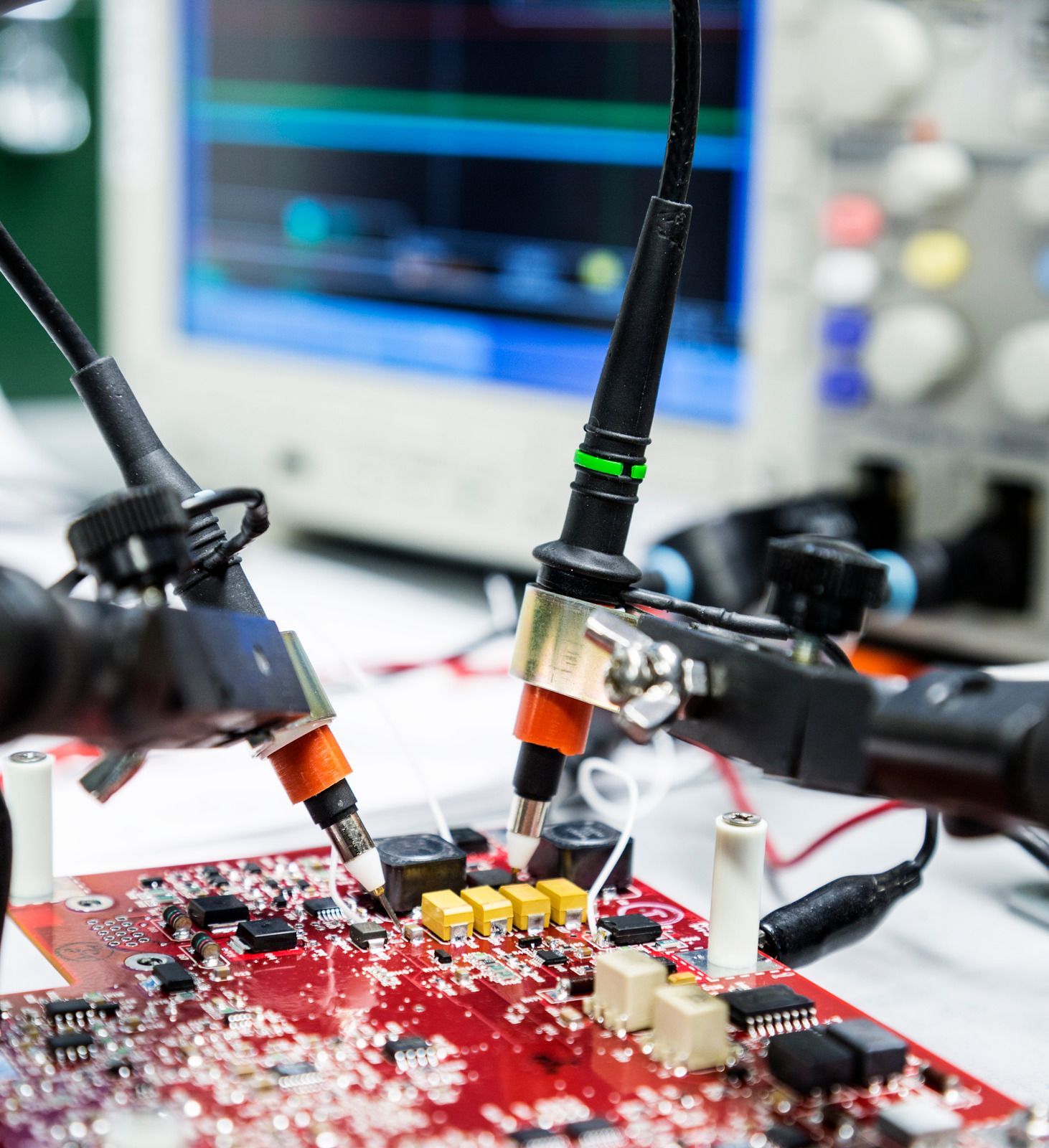 Circuit board with probes connected, being tested in a laboratory setting, oscilloscope in background.