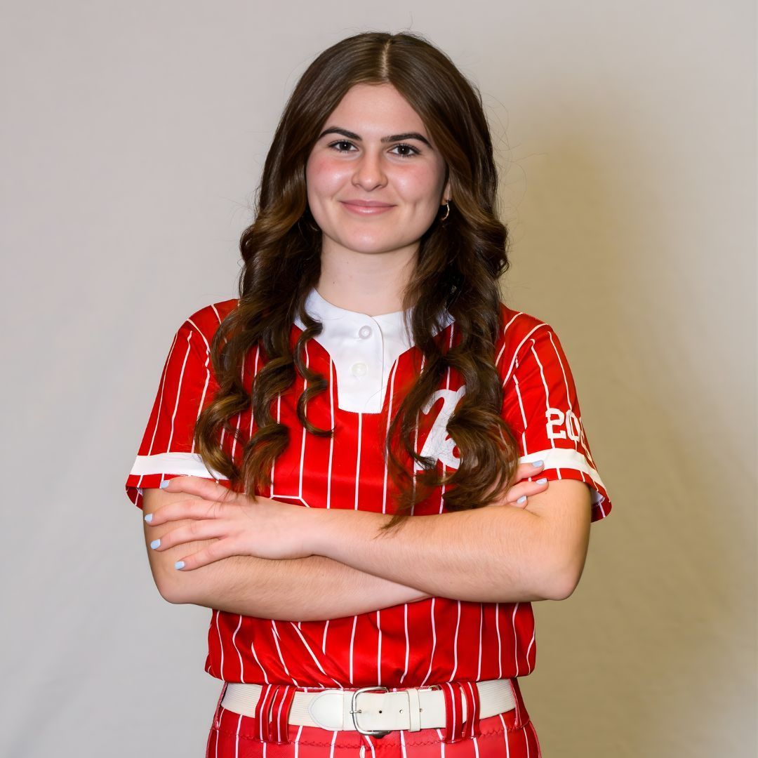 Smiling person with long hair wearing a red and white pinstriped softball jersey with arms crossed against a grey wall.