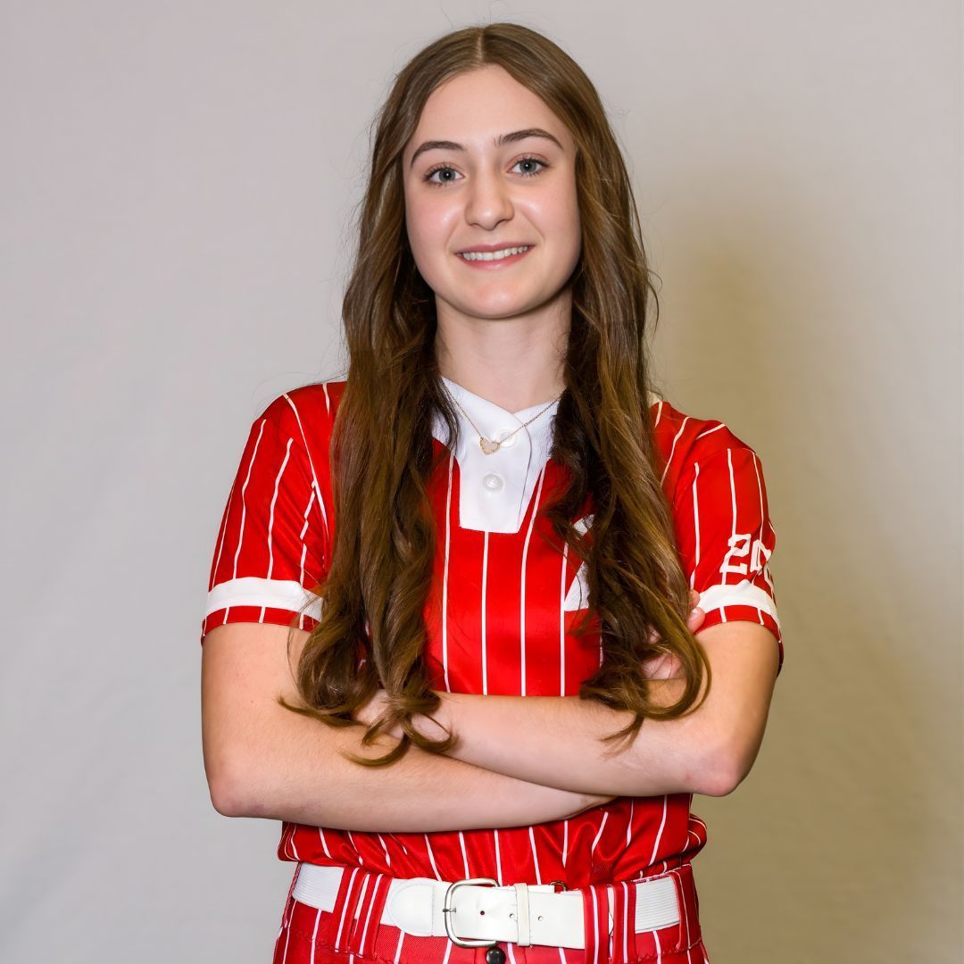 A person with long brown hair, smiling with arms crossed, wearing a red and white pinstriped baseball jersey and belt.