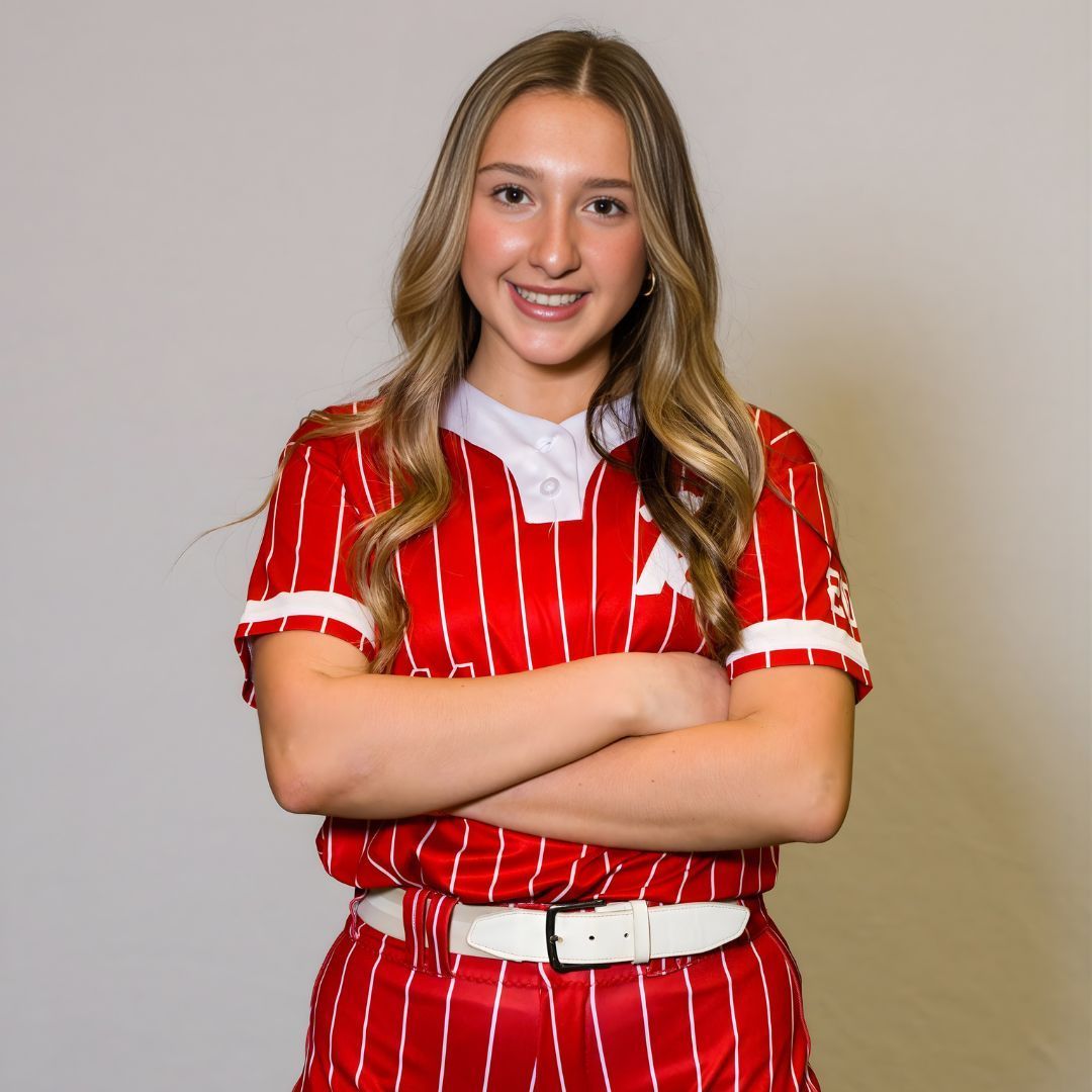 A person with long, blonde hair smiles with arms crossed, wearing a red and white pinstriped athletic uniform.