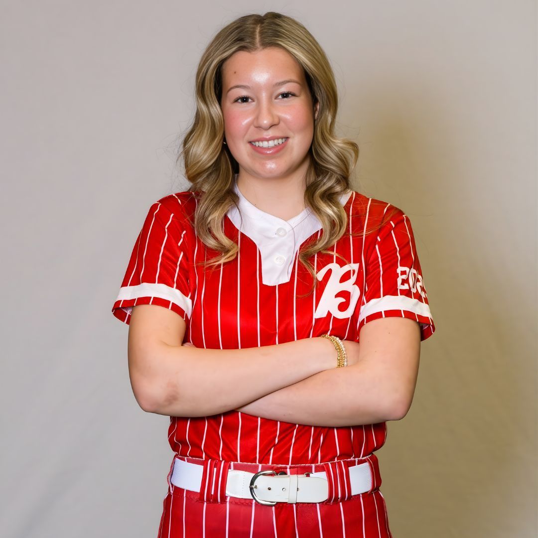 A smiling person with shoulder-length blonde hair, wearing a red and white pinstriped baseball jersey with arms crossed.