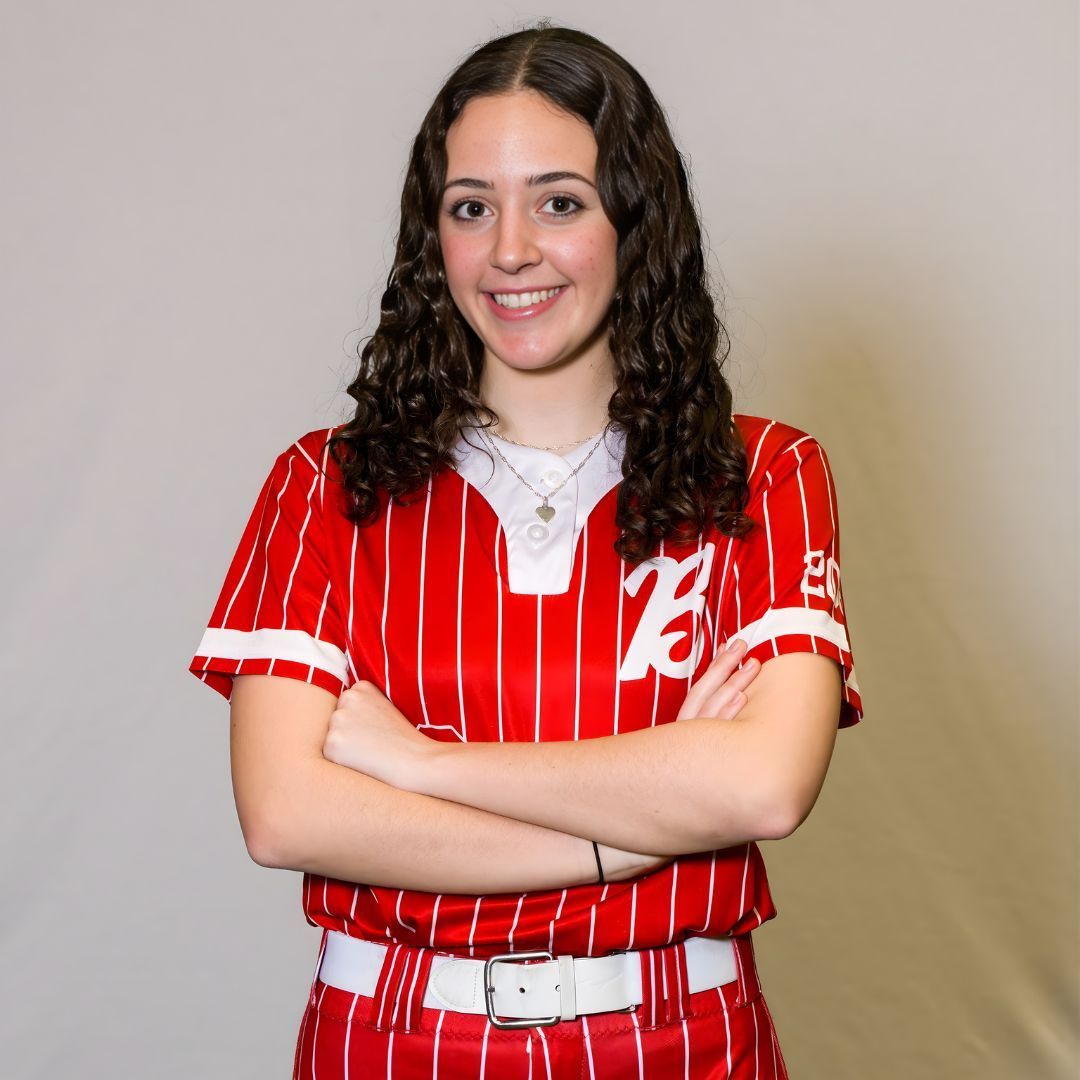 A person with long curly brown hair, wearing a red and white pinstriped softball uniform, stands with arms crossed.