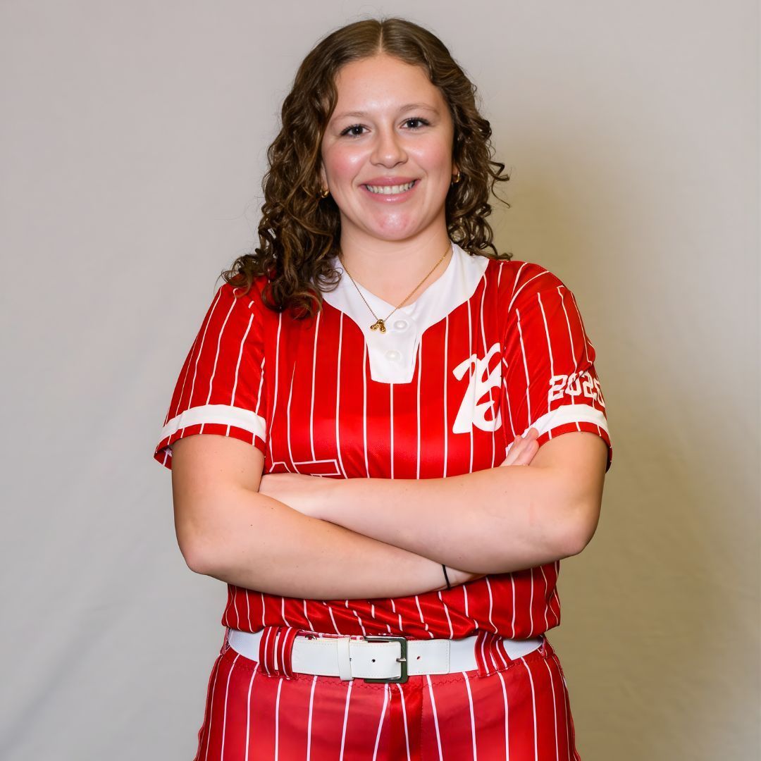 A person with curly brown hair smiling with arms crossed, wearing a red and white pinstriped baseball uniform.