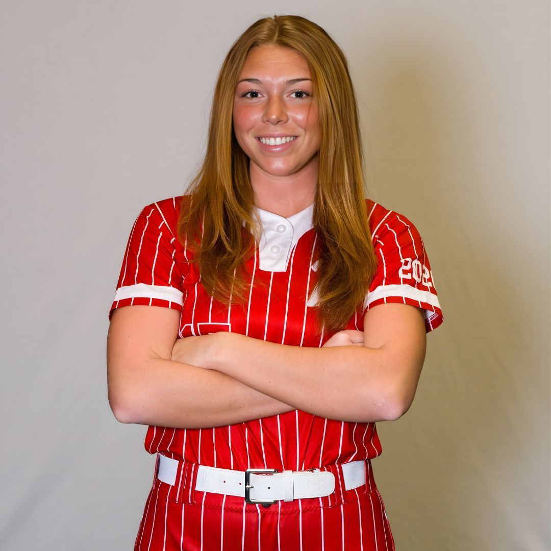 A smiling person with long hair wearing a red and white pinstriped softball uniform, arms crossed, against a gray background.