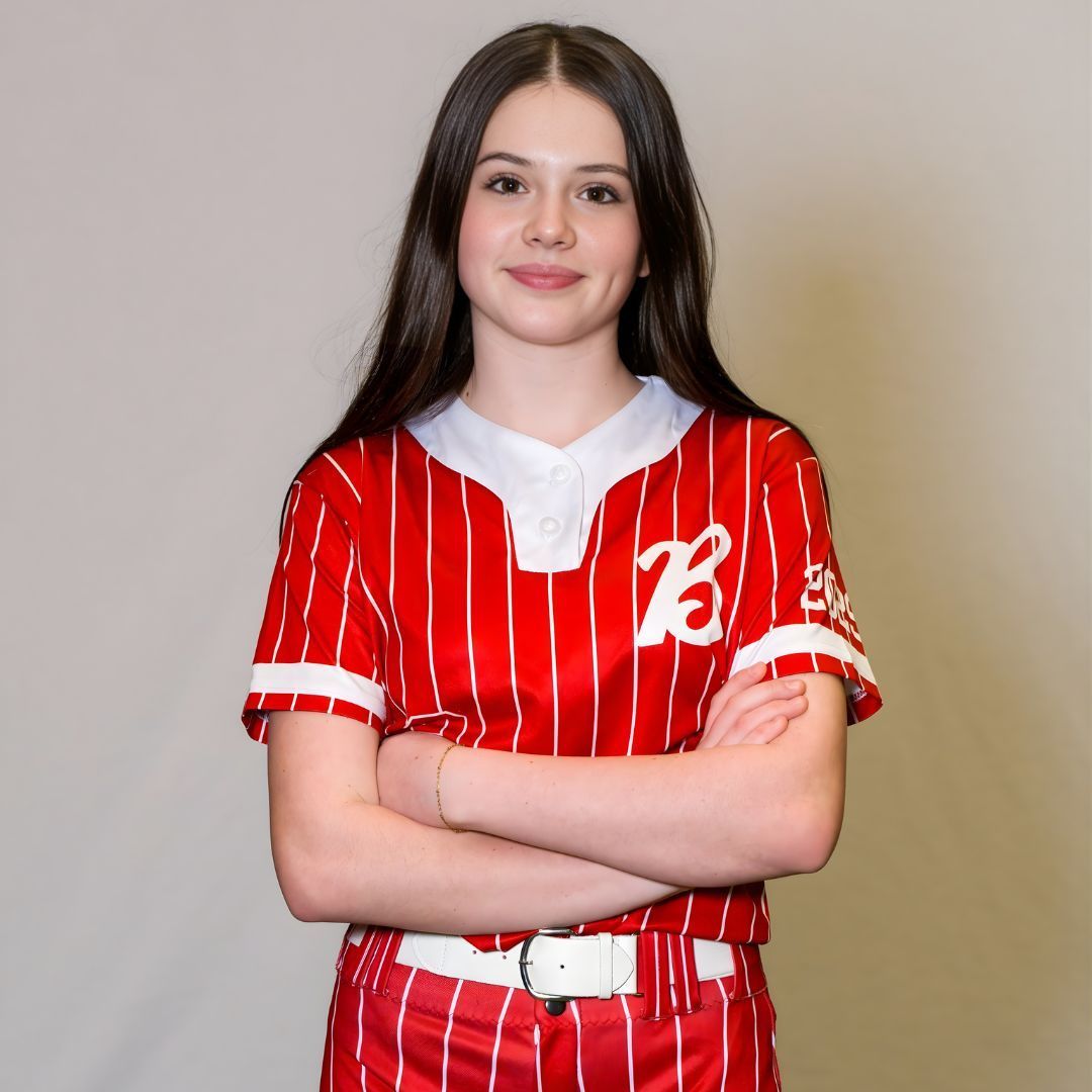 A person with long dark hair, wearing a red and white striped baseball jersey, stands with arms crossed against a backdrop.