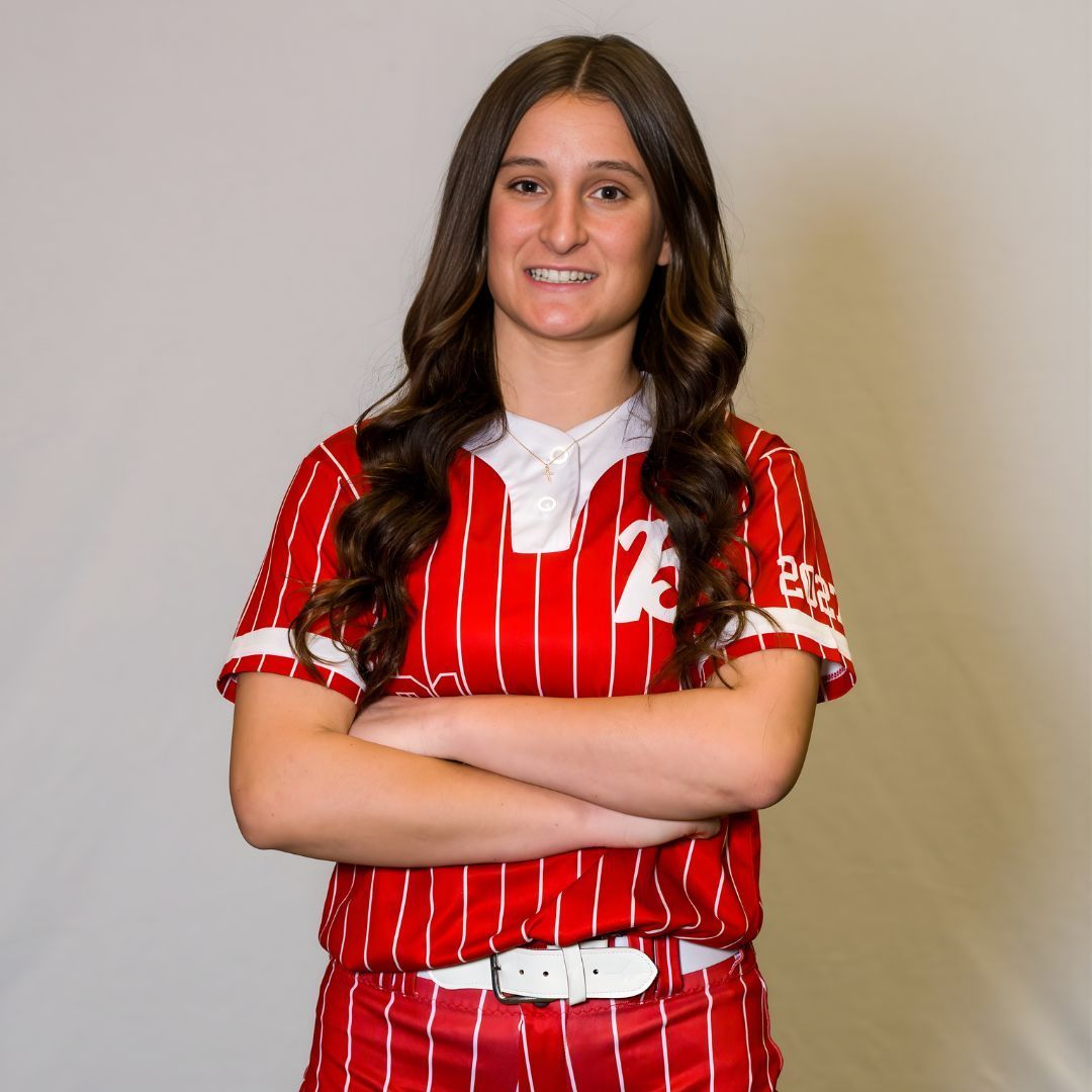 A person with long brown hair wearing a red and white striped softball uniform with arms crossed, smiling at the camera.