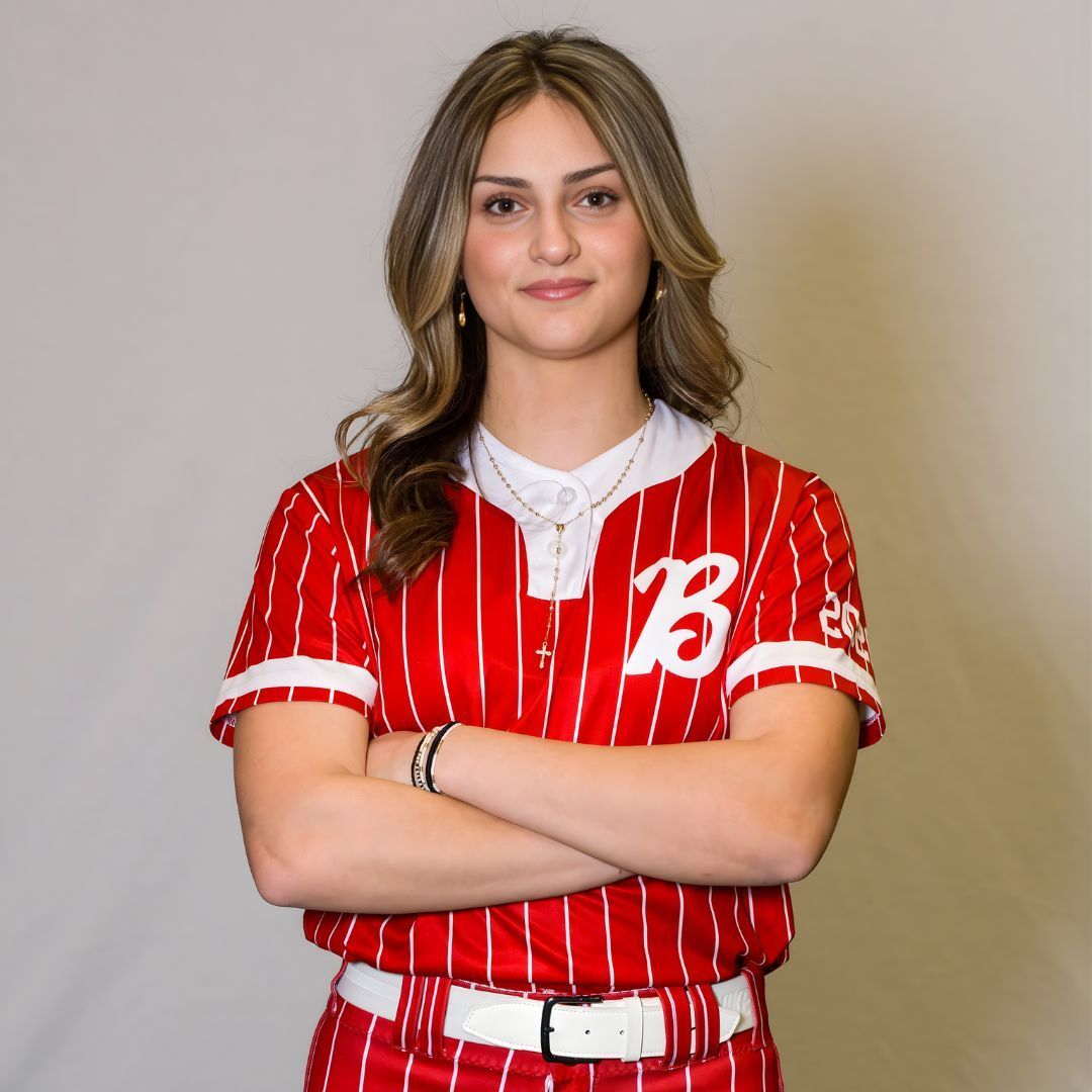 A person wears a red-and-white pinstriped softball jersey with a letter B, standing with arms crossed against a grey wall.