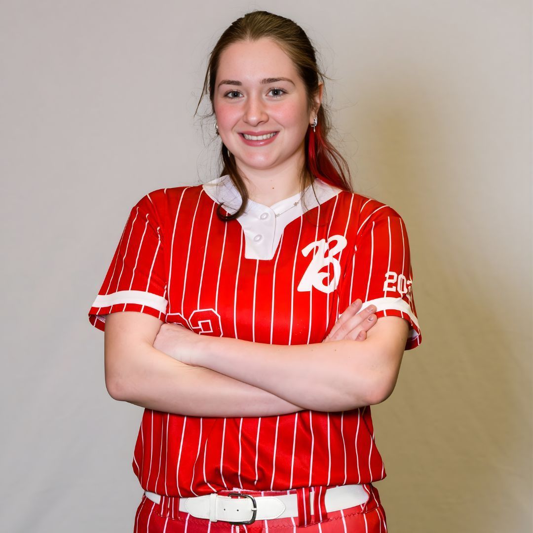 A smiling athlete wears a red-and-white pinstriped softball uniform with arms crossed against a plain grey background.