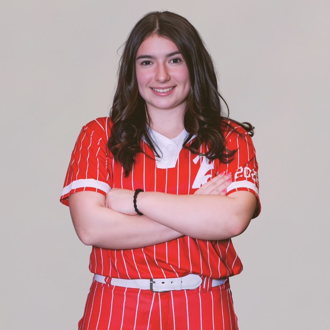 A smiling individual with long brown hair wearing a red and white striped baseball jersey with arms crossed.