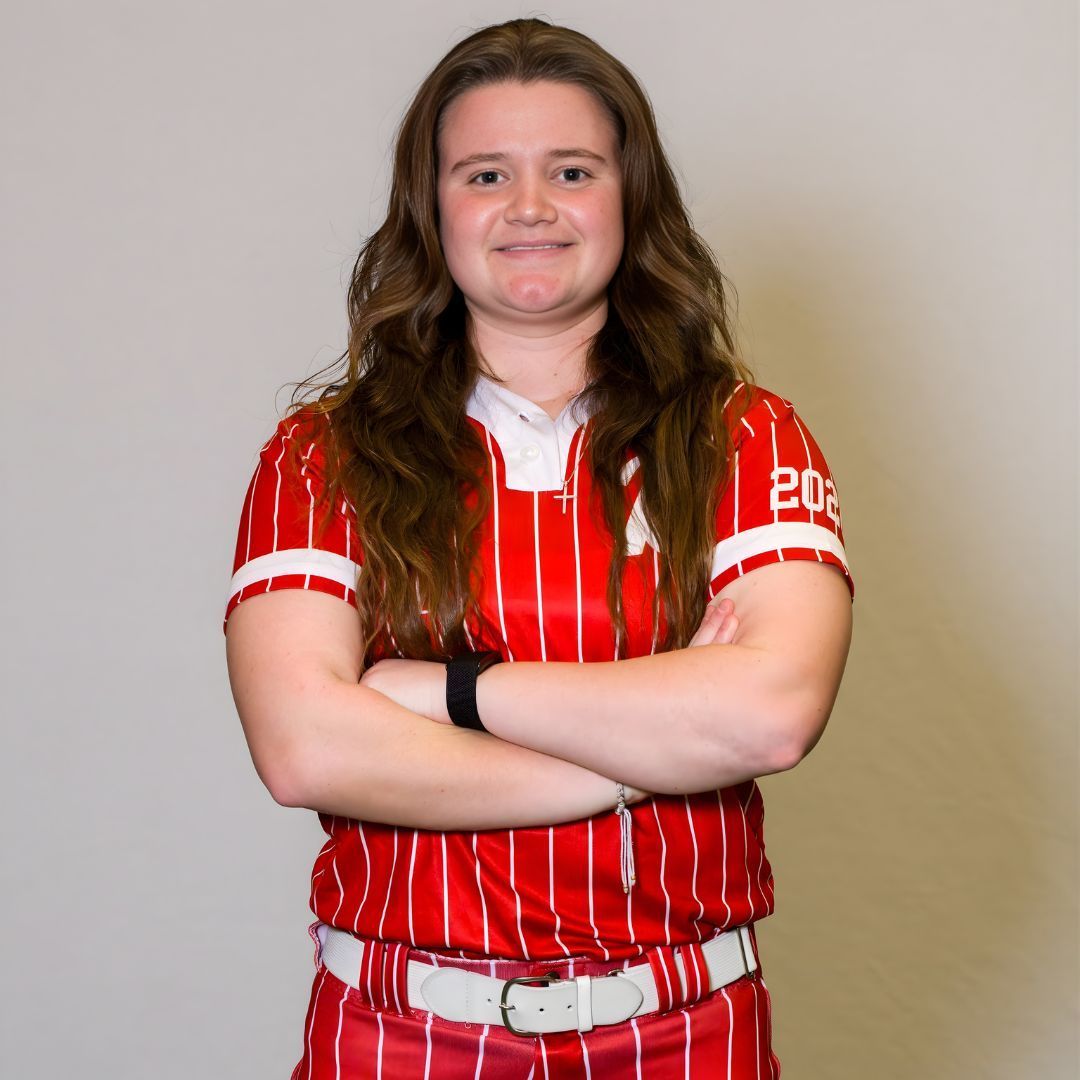 A person with long brown hair, wearing a red and white striped athletic uniform, stands with their arms crossed.