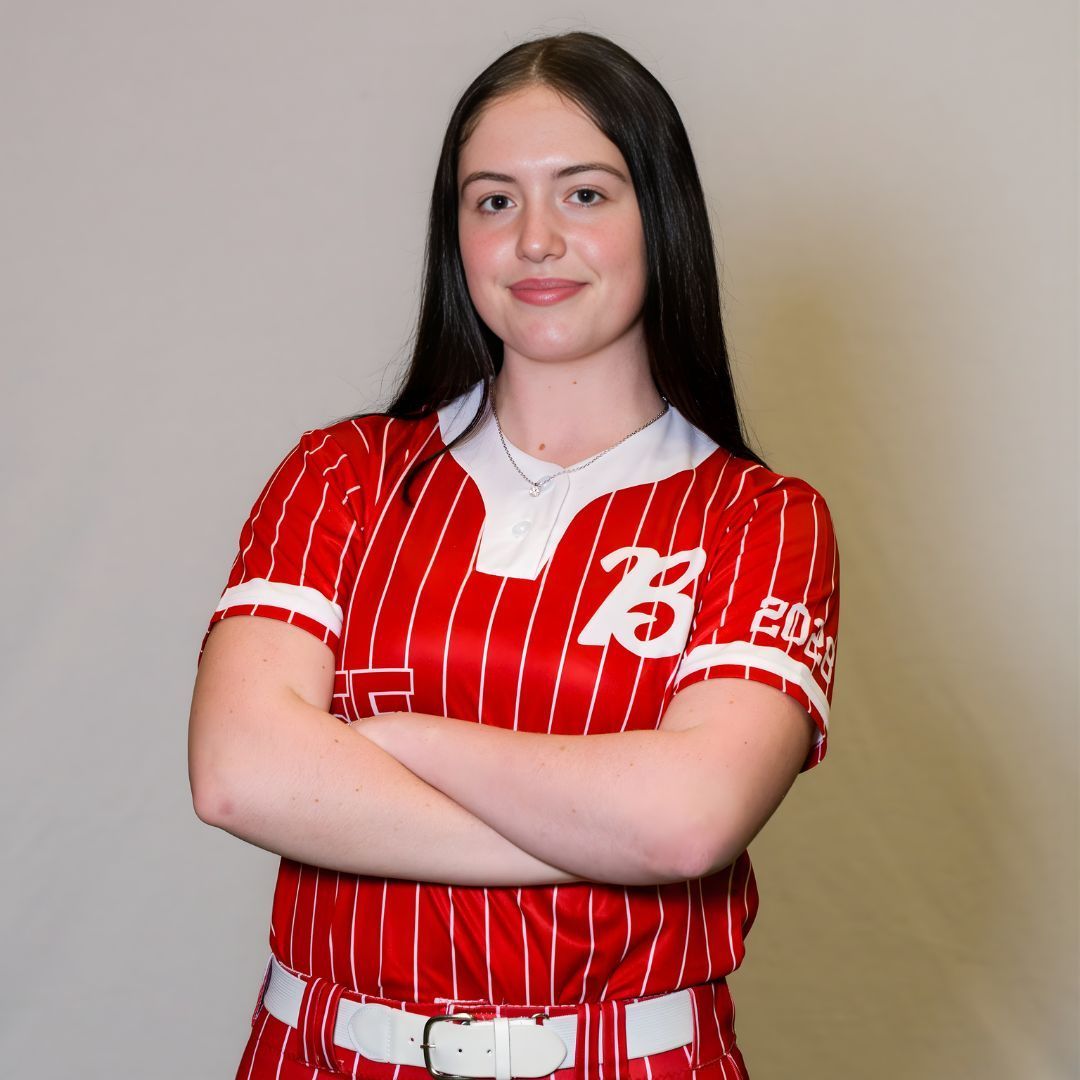 A person with long dark hair, wearing a red and white striped baseball jersey, stands with arms crossed against a backdrop.