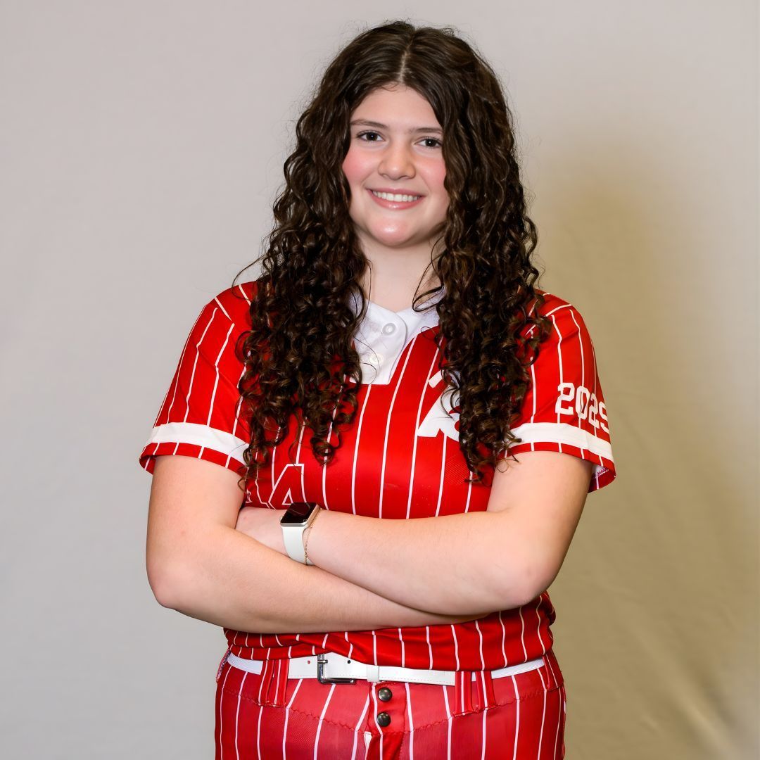 A person with long, curly dark hair smiles, arms crossed, wearing a red-and-white pinstriped softball uniform.