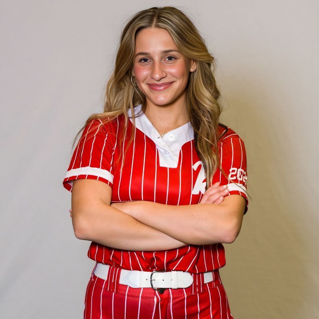 A person smiling with arms crossed, wearing a red and white pinstriped baseball jersey and belt against a plain background.