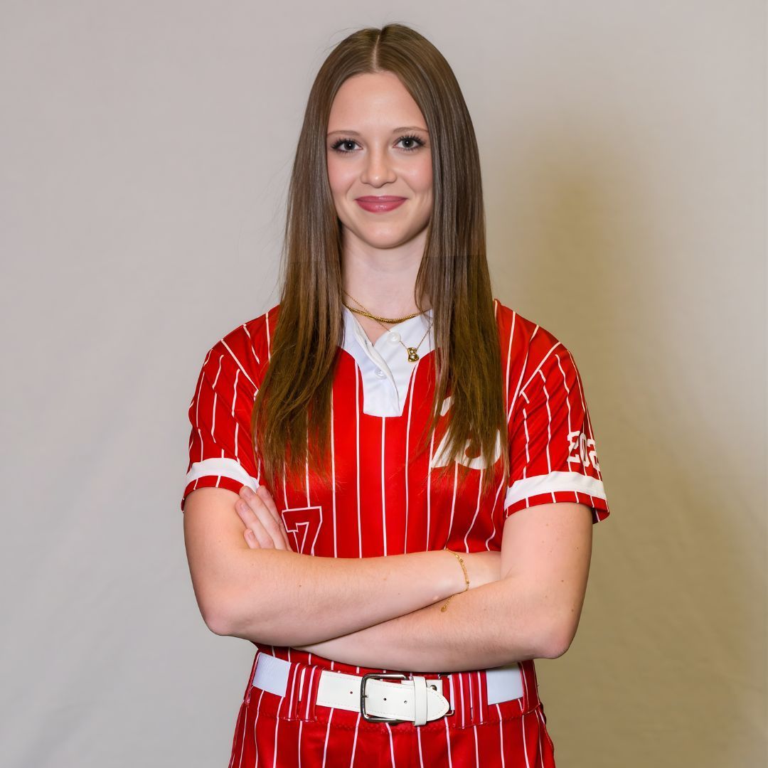 A person stands with arms crossed, wearing a red and white pinstriped sports jersey and white belt against a gray background.