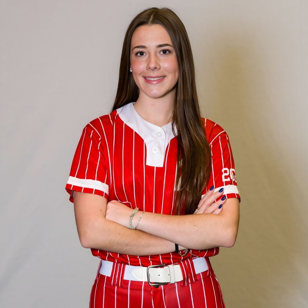 A smiling softball player in a red pinstriped uniform with arms crossed, set against a plain gray background.