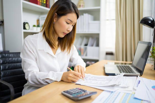 Woman in white shirt working at a desk with laptop, calculator, and papers, in an office.