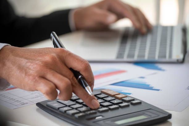 Person's hands using a calculator, pen, and laptop on a desk with financial charts.