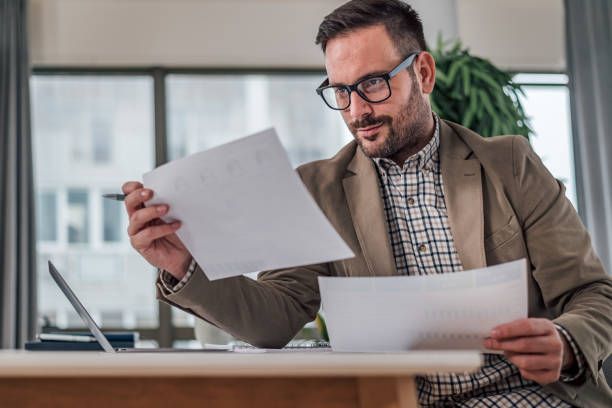 Man in glasses reviewing paperwork at desk near a window, with a laptop.